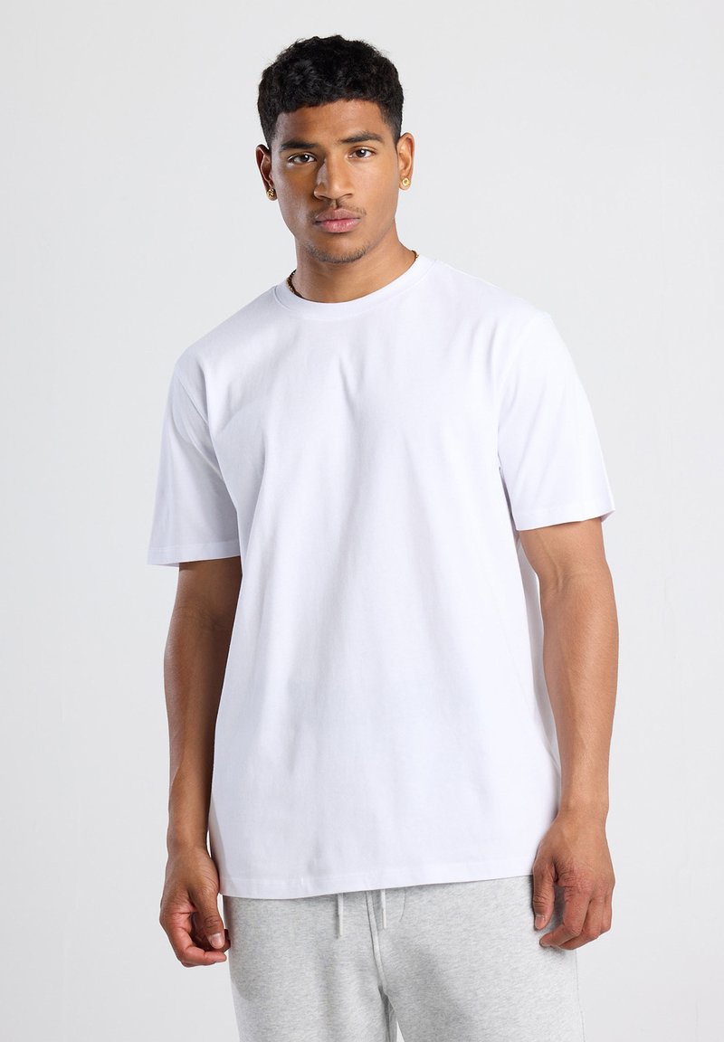 T-shirt en coton blanc à manches courtes, avec un col rond et une coupe standard, porté avec un pantalon de survêtement gris. Texture lisse sans motifs.