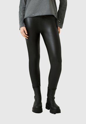 Person trägt schwarze Lederleggings, schwarze grobe Stiefel und ein langärmeliges graues Oberteil mit kleinen dekorativen Nieten an den Ärmeln.