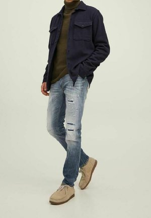 Mand iført en mørkeblå jakke, olivengrøn rullekrave, slidte blå jeans og beige ruskindssko står med den ene hånd i lommen.