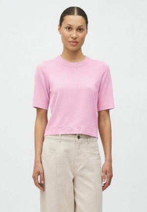 Frau mit zurückgebundenem dunklem Haar, trägt ein kurzärmeliges, rosafarbenes Strickoberteil und hoch taillierte beige Hosen vor einem schlichten Hintergrund.