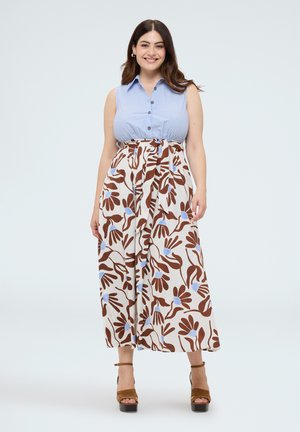 Femme souriante portant un chemisier rayé bleu sans manches et une jupe blanche avec un grand motif floral marron et bleu, debout dans des sandales à plateforme marron.