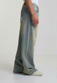 Denim-farkut vaaleansinisessä gradienissa, joissa on leveät lahkeet, kaksi takataskua logo-patchillä ja kontrastivärillä ommeltuna. Sileä rakenne.