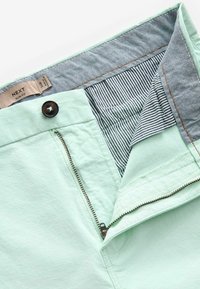 Shorts verde mentă cu fermoar, căptușeală interioară cu dungi albastre deschise și închidere cu nasture maro. Materialul pare moale, cu o ușoară textură.