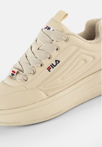 Beige sportcipő, textúrált szintetikus felsőrészel, fűzős záródással és logóval. Vastag talppal és jellegzetes oldalsó panel dizájnnal rendelkezik.