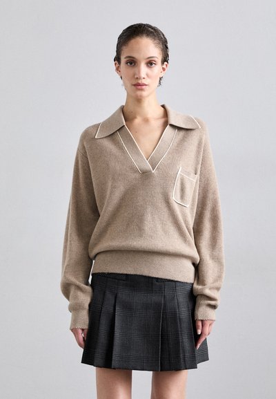 Pull en tricot beige avec un col en V et un piping blanc, associé à une jupe plissée à carreaux noirs. L'ensemble possède des manches longues et une poche poitrine.