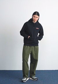 Felpa nera con un piccolo logo rosso, abbinata a pantaloni larghi verde oliva e sneakers grigie con dettagli neri, su un tappeto blu.