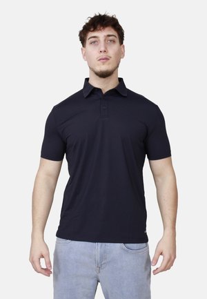 Giovane uomo in piedi rivolto verso l'obiettivo, indossa una polo nera a maniche corte e un paio di jeans azzurri, su sfondo bianco.