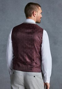 Gilet bordeaux à motif avec une finition texturée, porté avec une chemise blanche. L'arrière présente une bande et une boucle pour l'ajustement.