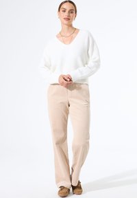 Vrouw staat in een witte V-hals trui, beige corduroy broek, bruine moccasinschoenen en gouden sieraden tegen een effen witte achtergrond.