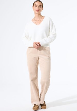 Mujer de pie vistiendo un suéter blanco de cuello en V, pantalones de pana beige, zapatos mocasines marrones y joyas doradas contra un fondo blanco liso.