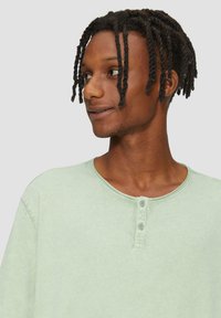 Jasnozielona koszula henley z długim rękawem, o okrągłym dekolcie, z dwoma guzikami przy kołnierzu oraz miękkim, teksturowanym materiałem.