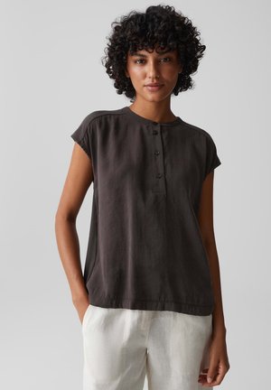 Femme aux cheveux bouclés portant un chemisier sombre à manches courtes boutonné et un pantalon clair, debout devant un fond gris uni.