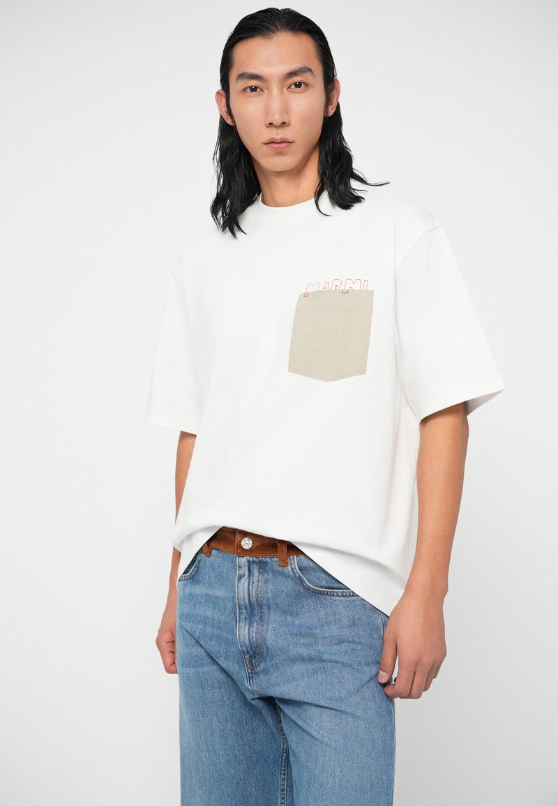 Junger Mann mit schulterlangem schwarzen Haar, der ein weißes T-Shirt mit einer beigen Tasche und blaue Jeans trägt, vor einem schlichten Hintergrund.