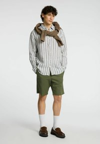 Camicia a righe bianca con linee multicolori; pantaloni corti verde oliva; scarpe marroni; maglione beige legato attorno al collo; calzini bianchi.