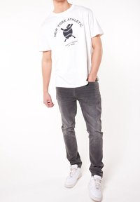 Camiseta blanca con un diseño gráfico negro que presenta una pelota de béisbol y bates cruzados, combinada con jeans grises y zapatillas blancas.