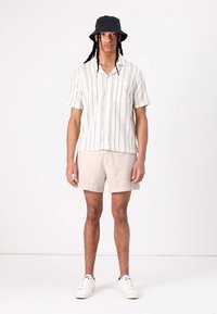 Korte mouwen gestreept shirt in crème met blauwe strepen, beige shorts en een zwarte bucket hat; witte sneakers met veters.