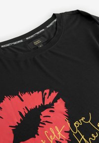 Must musta särk mustast puuvillast, millel on punane musi print ja kuldne tekst, ümar kaelus ja lühikesed varrukad. Etikett kannab nime "ROCKETT ST GEORGE."