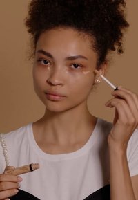 Tube de correcteur KIKO tenu dans une main, couleur beige, avec un applicateur éponge. Le modèle l'applique sous ses yeux, portant une chemise blanche et noire.