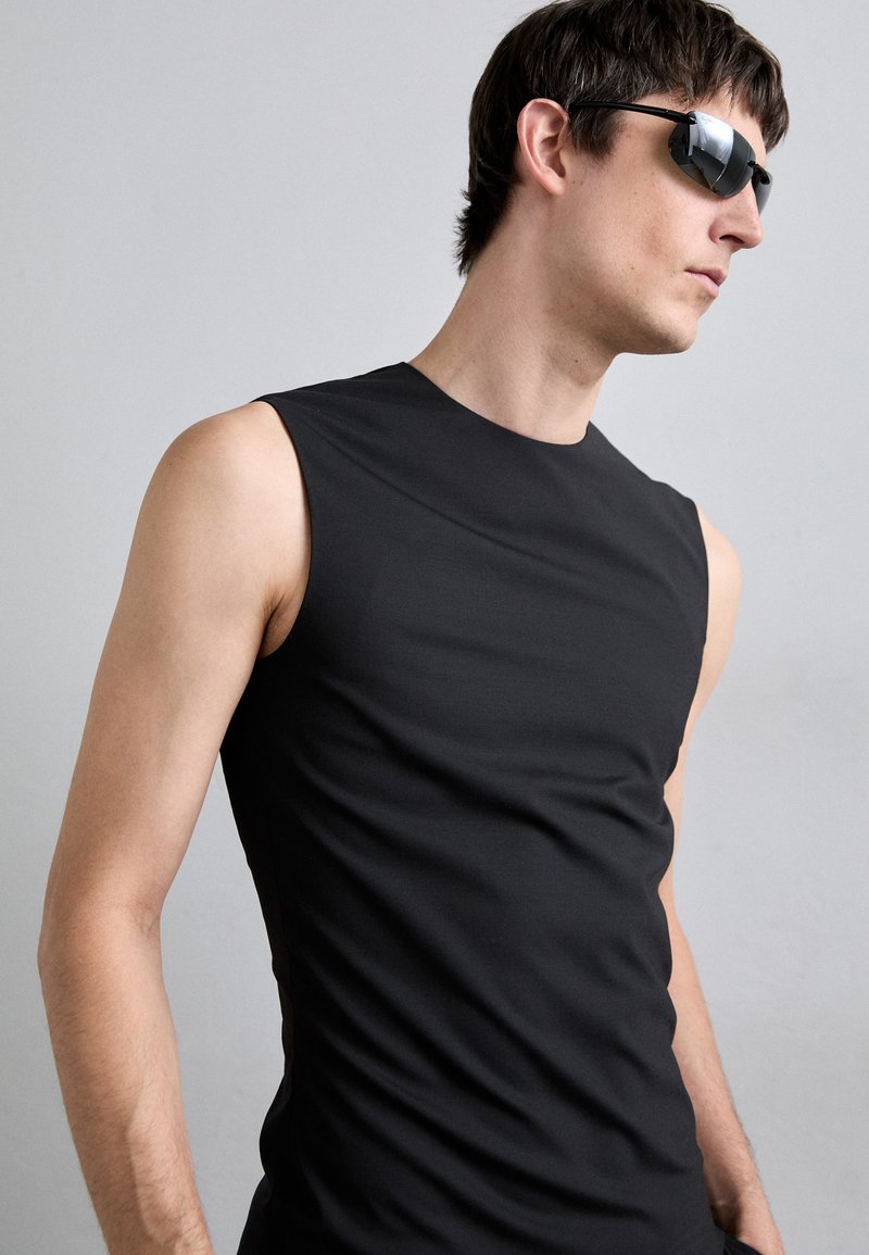Sleeveless black top, smooth fabric, fitted design, round neckline, minimal seams, worn with sunglasses.
