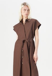 Brązowa sukienka do kolan z krótkimi rękawami, na guziki z przodu, z paskiem do wiązania. Wykonana z gładkiego materiału, o luźnym kroju i minimalistycznym designie.