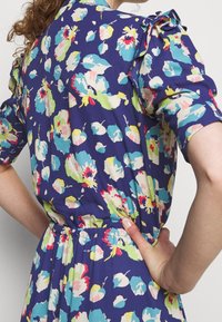 Květinové šaty z tmavomodrého materiálu s krátkými nabíranými rukávy, zdobené pestrými květinami v růžové, zelené a modré barvě, a elastickým pasem.