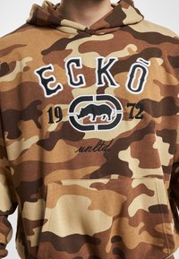 Brun camouflage hoodie med en frontlomme. Inkluderer broderet "ECKŌ" og "1972" tekst samt et grafik med en sort dyresilhuet.