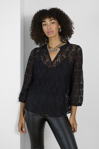 Schwarze Spitzenbluse mit V-Ausschnitt und Dreiviertelärmeln, verziert mit floralen Mustern. Kombiniert mit schwarzen, figurbetonten, glänzenden Hosen.