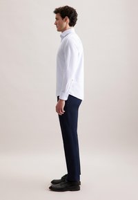 Camicia bianca a maniche lunghe con righe verticali, abbinata a pantaloni blu scuro e scarpe nere. La vista laterale mostra la vestibilità e i dettagli del polsino abbottonato.