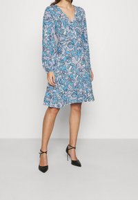Blumenkleid mit langen Ärmeln, das über einen V-Ausschnitt verfügt. Blau-graues Muster auf hellem Hintergrund. Kombiniert mit schwarzen High-Heels.
