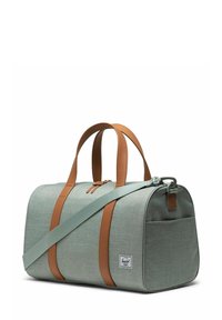 Sac de sport en tissu vert clair avec des accents en cuir marron. Il dispose de deux poignées et d'une bandoulière réglable. Patch de logo visible.