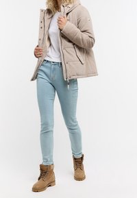 Beżowa kurtka puchowa z korduroy z kapturem, jasnoniebieskie dopasowane dżinsy oraz jasnobrązowe buty trekkingowe z wiązanym detalem.