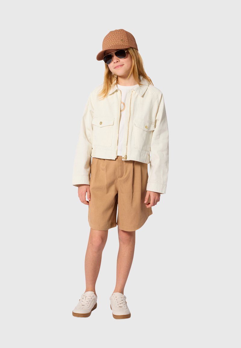 Jeune personne portant une casquette beige, des lunettes de soleil foncées, une veste blanche, une chemise blanche, un short marron et des baskets blanches, debout devant un fond gris.