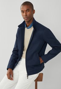 Marineblå zip-up jakke med høj krave, båret over en beige sweater og denimskjorte, har en glat tekstur og minimalistisk design.