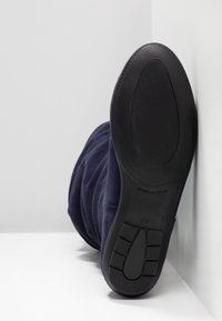 Semelle d'une botte bleu marine appuyée contre un mur blanc, montrant une semelle extérieure en caoutchouc noir texturée avec la taille 37 et l'inscription « Fabriqué en Espagne ».