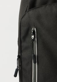 Λεπτομέρεια μαύρου υφασμάτινου σακιδίου πλάτης που διαθέτει τσέπη με φερμουάρ και ταινία έλξης, αντίθετη γκρι ραφή και υφή με ανάγλυφο φινίρισμα.