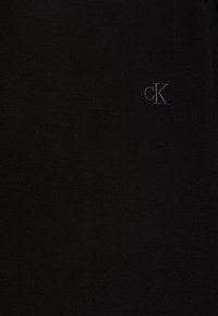 T-shirt preto de algodão com uma textura suave, apresentando um pequeno logótipo "cK" bordado em uma tonalidade mais escura à esquerda.