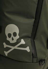 Mochila verde oliva con un gran diseño de calavera y huesos cruzados en gris, que presenta un tejido texturizado, un bolsillo con cremallera y detalles en negro.