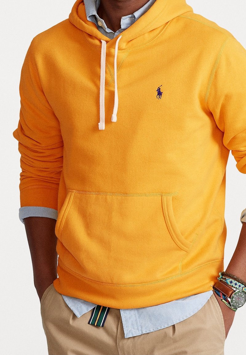 Orange hoodie med vita dragsko och en kenguruficka; har en liten blå logotyp på bröstet. Kombinerad med en ljusblå skjorta med krage.