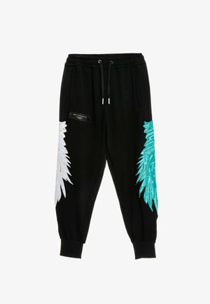 Joven está de pie, vistiendo una sudadera con capucha negra y pantalones con diseños de alas blancas y turquesas, camiseta blanca y botas blancas con cordones.