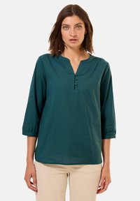 Blusa teal con maniche a tre quarti, scollatura a V e tre bottoni scuri sul davanti. Tessuto morbido, vestibilità casual e leggero arricciamento alle maniche.