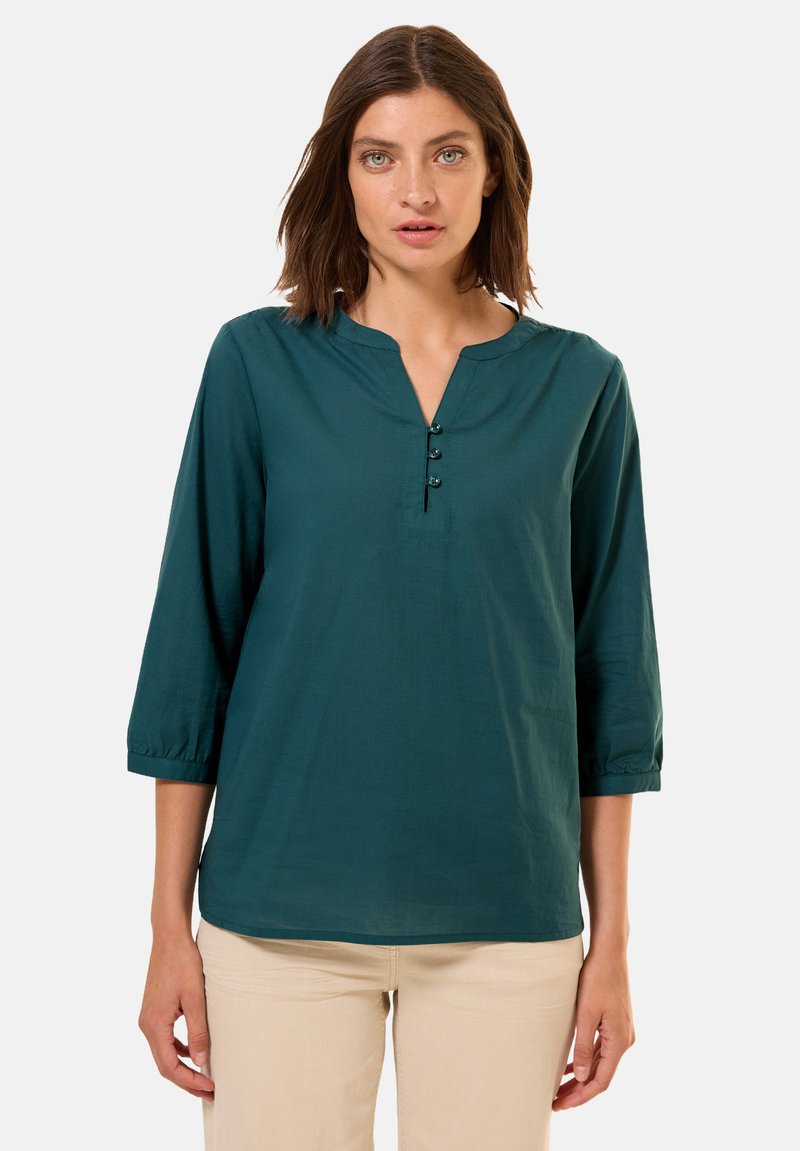 Blusa teal con maniche a tre quarti, scollatura a V e tre bottoni scuri sul davanti. Tessuto morbido, vestibilità casual e leggero arricciamento alle maniche.