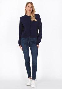 Merino-neule, jossa on pyöreä pääntie, pitkät hihat ja nappikiinnitys, yhdistettynä slim fit -tummaan farkkuun ja valkoisiin lenkkareihin.