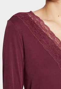 Burgunderfarbener Oberteil mit gerippter Textur, versehen mit einem tiefen V-Ausschnitt, der mit einer Spitzenkante entlang des Kragens geschmückt ist. Lange Ärmel sichtbar.