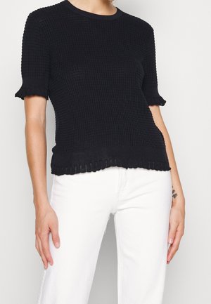Femme portant un haut noir texturé à manches courtes avec des bords froncés, associé à un pantalon blanc taille haute, debout devant un fond uni.
