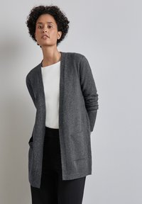 Cardigan grigio aperto realizzato in morbido tessuto a maglia; presenta polsini a costine e due tasche frontali, indossato sopra una maglia bianca.