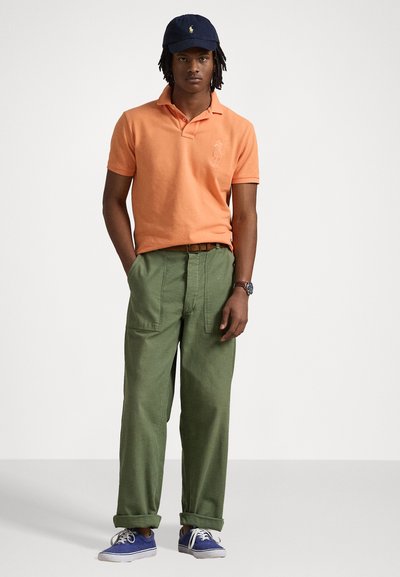 Oranje poloshirt, groene broek met een losse pasvorm, marineblauw pet en blauwe sneakers. Kenmerken zijn een logo op het shirt en een textuurstof.
