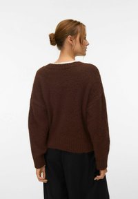 Femme aux cheveux châtain clair attachés en chignon, portant un pull en maille marron foncé ample et un pantalon noir, vue de dos sur un fond blanc uni.