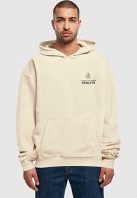 Beige hoodie met een kangoeroezak, voorzien van zwarte tekst "SPORT'S SEASON ATHLETE" op de borst. Zachte stoftextuur.