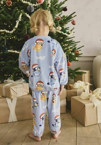 Vaaleansininen pyjama-asukokonaisuus, jossa on sarjakuvahahmoja joulupukilla. Pehmeää kangasta, joustavat hihansuut ja vyötärö. Taustalla joulukuusi.