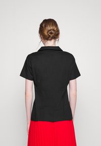 Camicia nera a maniche corte con colletto, design aderente e tessuto morbido; indossata con una gonna plissettata rossa.